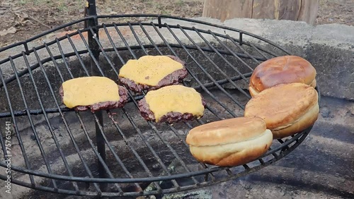 Wild game deer burgers with cheese melting on top being grilled over fire pit next to roasting buns on the side. photo