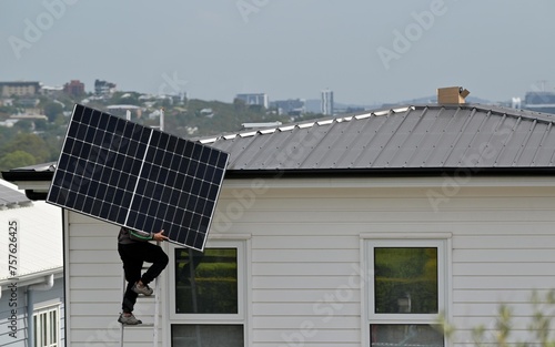 Solar construction worker  on a high ladder carry a large solar panel to a house roof