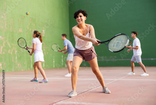 Sporty young Argentinian woman playing popular team match of frontenis at open-air fronton on summer day, ready to hit rubber ball with racquet © JackF
