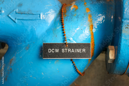 DCW Strainer label on pipe in water meter room. Close up of blue pipe with name tag and arrow to the domestic cold water strainer. Used to filter and remove particles from water. Selective focus. photo