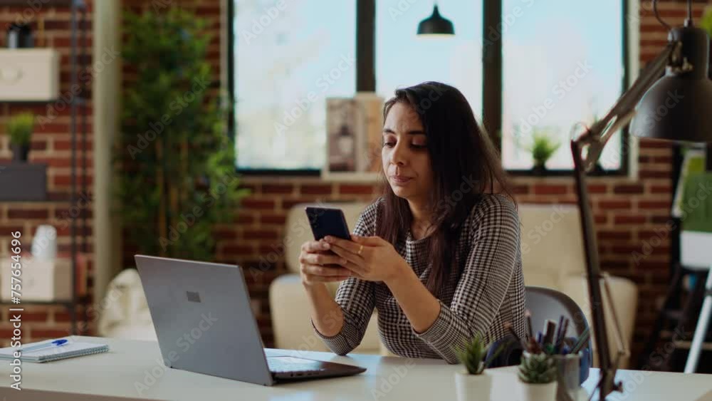 Teleworker checking her phone notifications at home in front of laptop, taking break from work. Woman in cozy apartment office taking pause from job tasks to read messages on smartphone, camera B Teleworker checking her phone notifications at home in front of laptop, taking break from work. Woman in cozy apartment office taking pause from job tasks to read messages on smartphone, camera B
