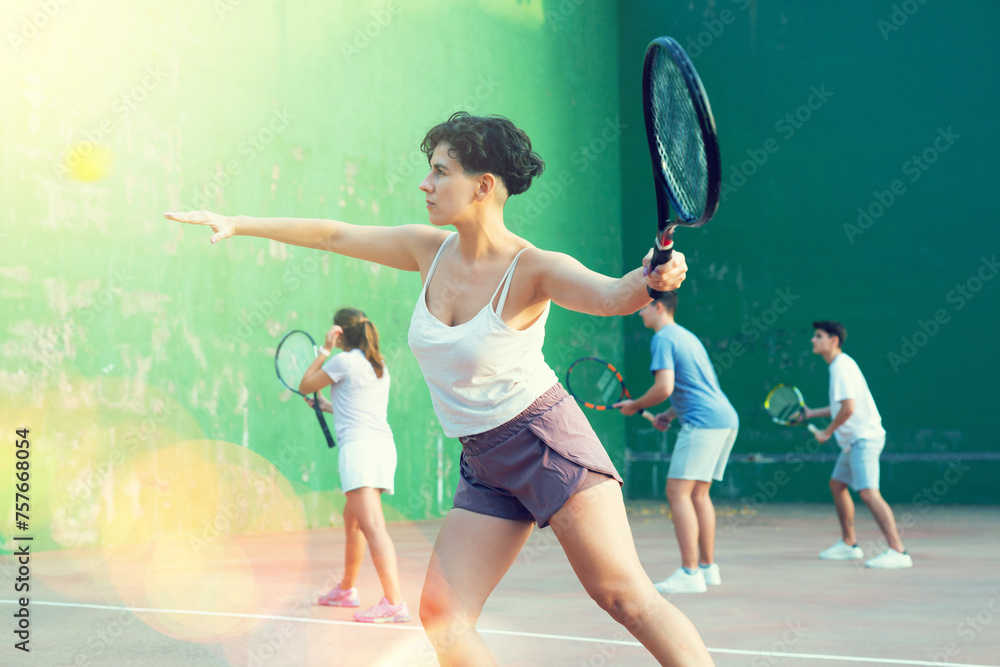 Sporty young Argentinian woman playing popular team match of frontenis at open-air fronton on summer day, ready to hit rubber ball with racquet