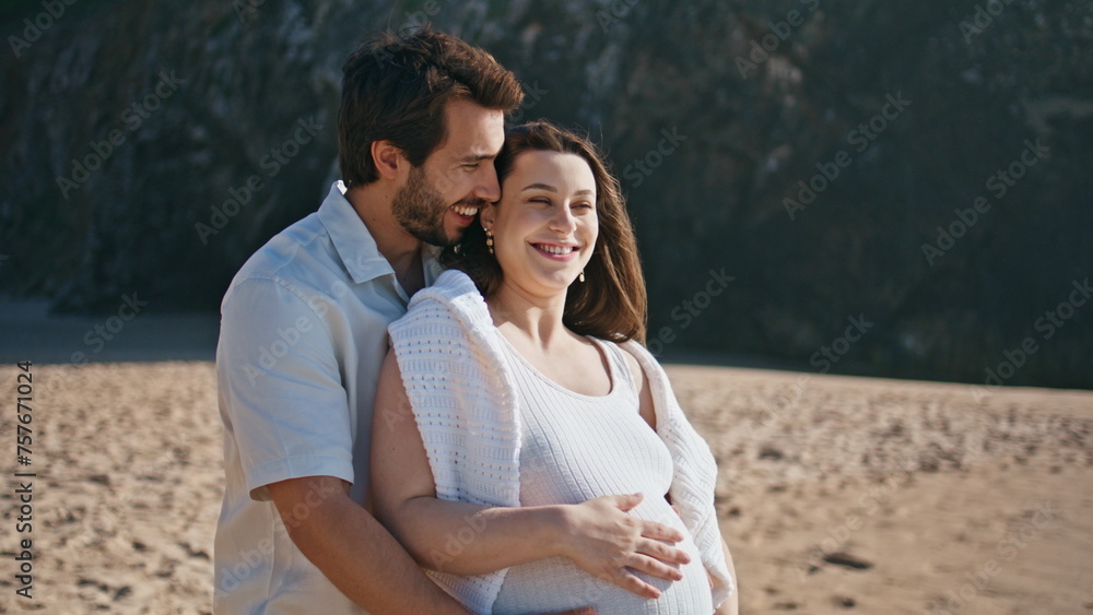 Obraz premium Smiling man embracing pregnant belly wife standing sandy seashore close up.