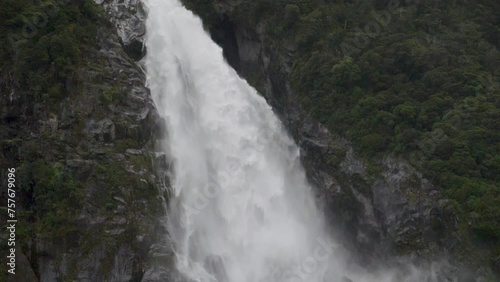 Slow Motion pan down a large waterfall on a cliff with a forest in foreground - Milford Sound (Piopiotahi), New Zealand photo