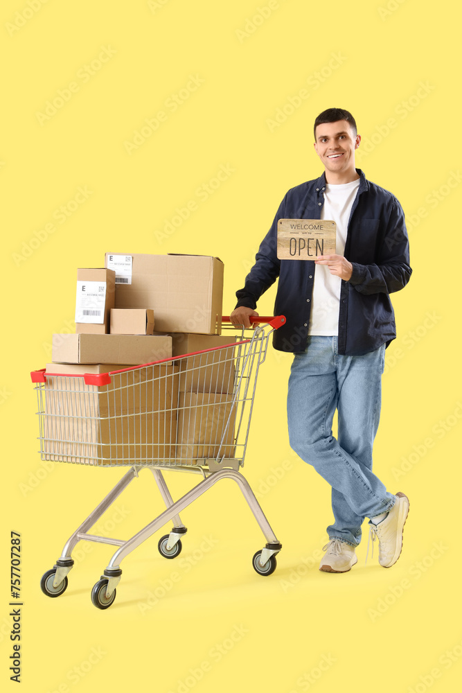 Male online store seller with OPEN sign and shopping cart of parcel boxes on yellow background Male online store seller with OPEN sign and shopping cart of parcel boxes on yellow background