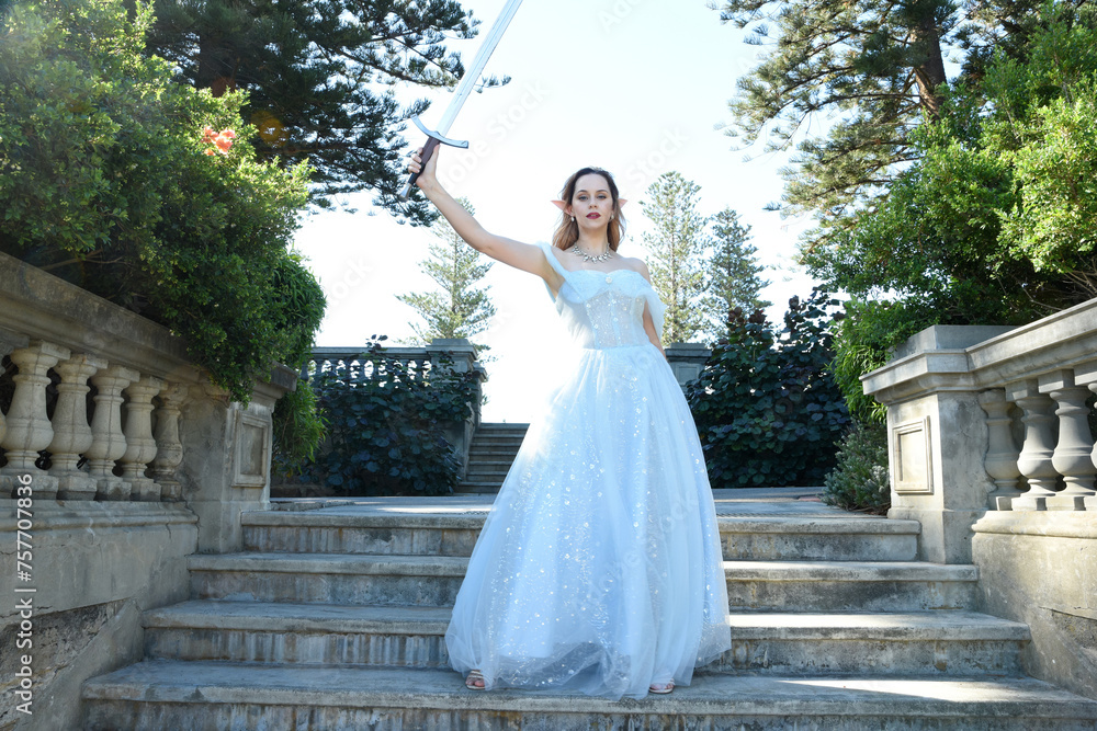 full length portrait of beautiful female model wearing blue fantasy ballgown, like a fairytale elf princess. Holding. Sword weapon, standing on staircase of a romantic castle balcony location.