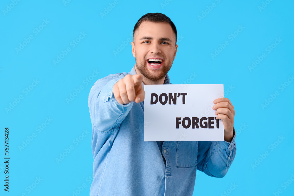 Young man holding paper with text DON'T FORGET and pointing at viewer on blue background Young man holding paper with text DON'T FORGET and pointing at viewer on blue background