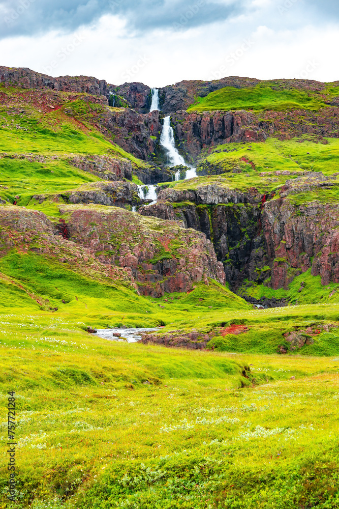Cover page with cascading waterfall in East Iceland, with dramatic sky and green meadow hills. Beautiful Icelandic landscape, huge cliff, water cascade and rock in moss