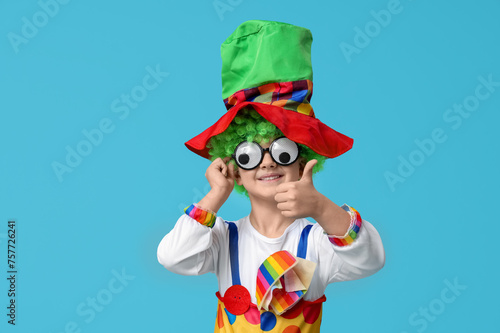Cute little boy in clown costume with funny glasses showing thumb-up on blue background. April Fools Day celebration