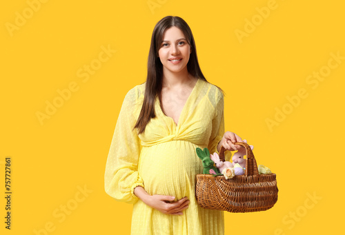 Young pregnant woman holding wicker basket with toys bunny and beautiful roses on yellow background. Easter celebration