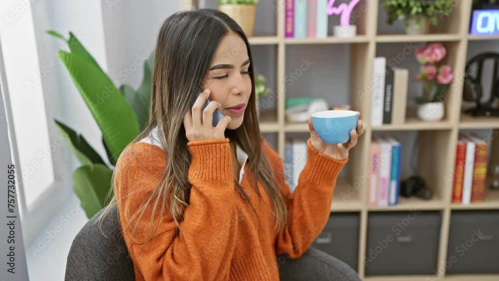 Young hispanic woman in an orange sweater talking on phone at home while holding a mug. Young hispanic woman in an orange sweater talking on phone at home while holding a mug.