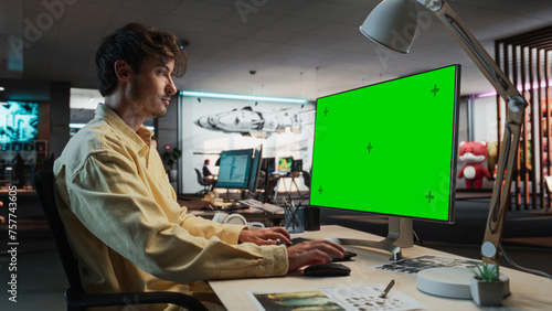 Caucasian Man Using Desktop Computer with Mock-up Green Screen Chromakey. Male Concept Artist Working in Game Design Startup Diverse Office, Creating Immersive Gameplay For New Survival Video Game.