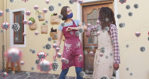 Image of covid 19 cells over two women in pottery studio wearing face masks greeting with elbows