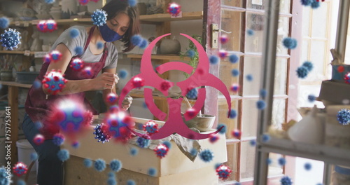Image of virus cells and biohazard symbol over caucasian woman with face mask forming pottery