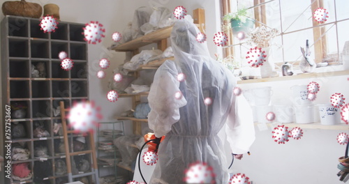 Image of covid 19 cells over person in ppe suit and face mask disinfecting pottery studio