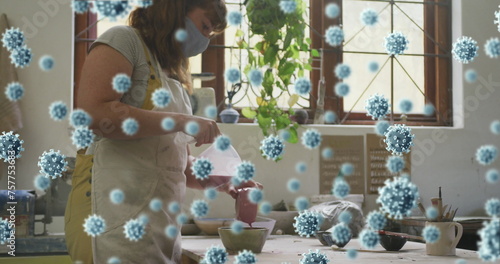 Image of covid 19 cells over woman working in pottery studio wearing face mask