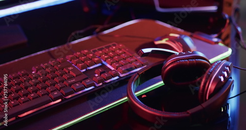 A gaming headset rests on a desk beside a keyboard, illuminated by neon lights