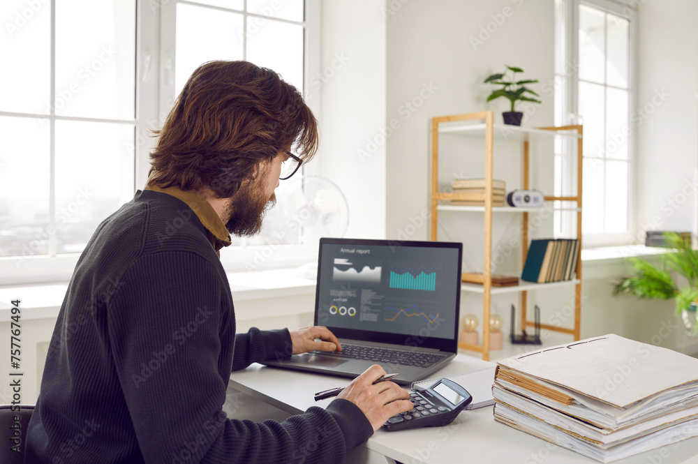 View over shoulder of busy man sitting at office desk, working with data, looking at graphs, charts and diagrams on screen of modern laptop computer, using calculator, and doing financial analysis View over shoulder of busy man sitting at office desk, working with data, looking at graphs, charts and diagrams on screen of modern laptop computer, using calculator, and doing financial analysis
