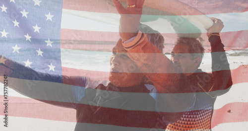 Image of flag of united states of america over senior biracial couple with surfboard on beach
