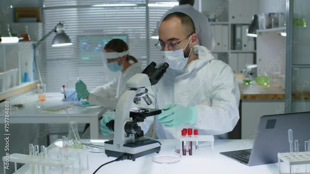Medium shot of Middle Eastern male scientist in protective coverall and face mask sitting at table and using microscope while working with diverse colleagues in modern laboratory Medium shot of Middle Eastern male scientist in protective coverall and face mask sitting at table and using microscope while working with diverse colleagues in modern laboratory
