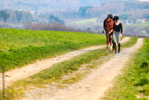 Horse and rider, saddled in riding clothes, walk side by side up a dirt road through green fields. © RD-Fotografie