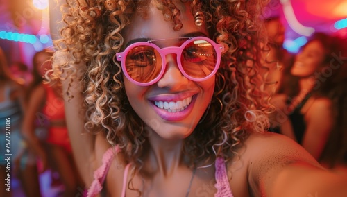Photo of an excited woman with big curly blonde hair and colorful taking selfie at the club party, surrounded by friends dancing around