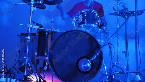 A drummer is captured in the midst of a performance on stage, surrounded by a drum kit with various cymbals and drumsticks in hand, under the bright lights of the venue.