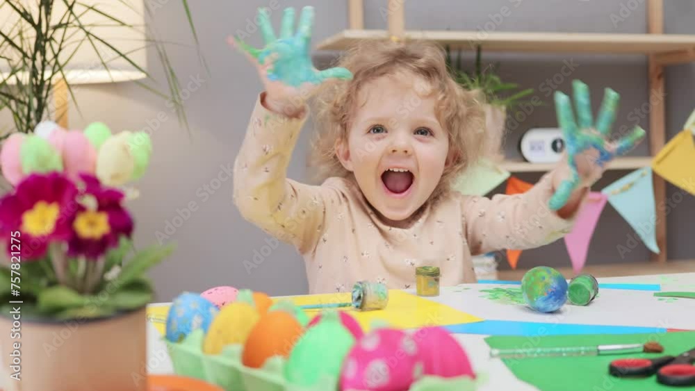 Easter egg painting fun. Creative egg coloring. Baby girl with curly blonde hair painting on Easter eggs at home showing her dirty colorful hands and having fun happily Easter egg painting fun. Creative egg coloring. Baby girl with curly blonde hair painting on Easter eggs at home showing her dirty colorful hands and having fun happily