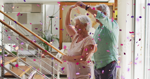 Image of pink and purple confetti falling over happy caucasian senior couple dancing at home