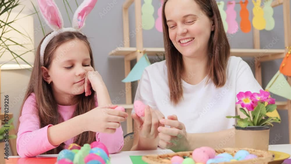 Delighted young adult woman with little daughter painting Easter eggs in festive home interior sitting together decorating holiday symbols with enthusiasm Delighted young adult woman with little daughter painting Easter eggs in festive home interior sitting together decorating holiday symbols with enthusiasm