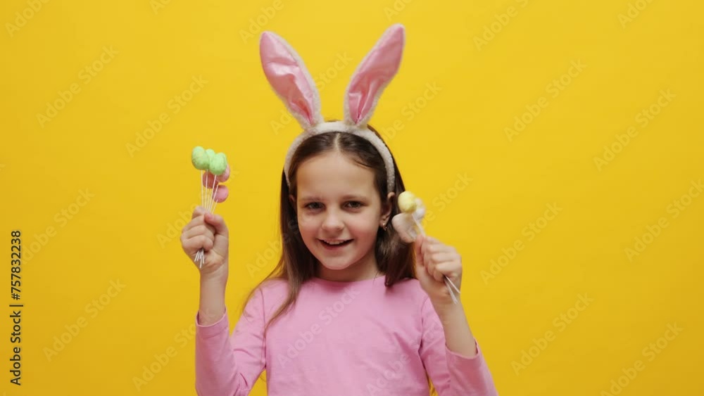 Child's joyful celebration of Easter. Overjoyed optimistic little girl wearing pink bunny ears headband holding pop cakes standing isolated over yellow background screaming exclaiming with excitement Child's joyful celebration of Easter. Overjoyed optimistic little girl wearing pink bunny ears headband holding pop cakes standing isolated over yellow background screaming exclaiming with excitement