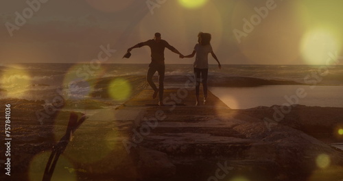 Yellow spots of light over african american couple holding hands walking on the rocks near the sea