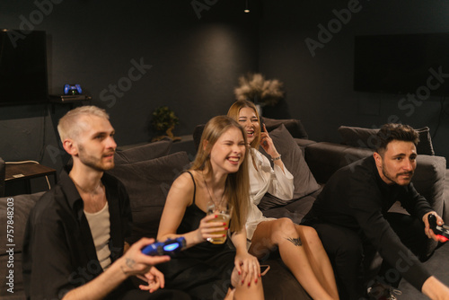 Young women laugh sitting between men playing console. Females share jovial banter with male counterparts enhancing vibrant atmosphere
