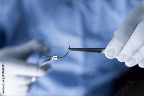 Closeup photo of a medical professional in blue scrubs handling sutures. Operator holding half circle needle with forceps and needle holder during operation.