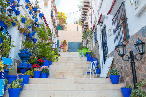 Narrow street with steps, white houses and blue potted plants in ancient neighborhood Santa Cruz in Alicante old town on hillside. Costa Blanca on Mediterranean sea coast, Spain