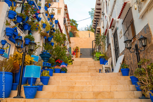 Narrow street with steps, white houses and blue potted plants in ancient neighborhood Santa Cruz in Alicante old town on hillside. Costa Blanca on Mediterranean sea coast, Spain © johnkruger1