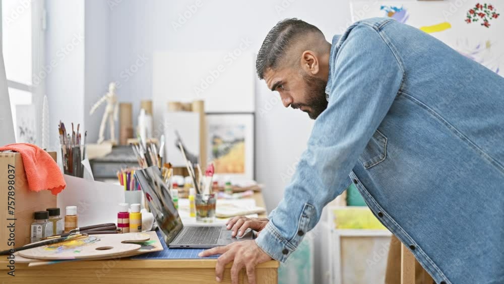 A focused bearded man using laptop in a bright art studio surrounded by painting supplies. A focused bearded man using laptop in a bright art studio surrounded by painting supplies.