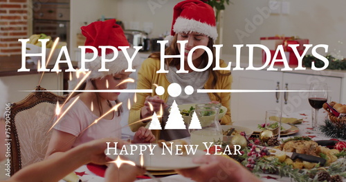 Image of happy holidays and new year text in white over family in santa hats at dinner table