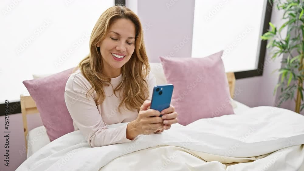 Smiling woman using smartphone in a cozy bedroom setting, portraying casual, relaxed home life. Smiling woman using smartphone in a cozy bedroom setting, portraying casual, relaxed home life.