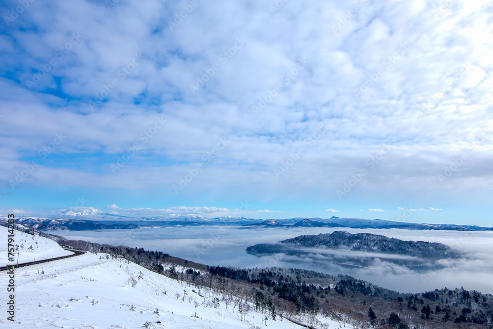 雪に覆われた山の斜面と眼下の湖の雄大な冬景色。雲の空の朝陽。北海道の美幌峠から見下ろす屈斜路湖。