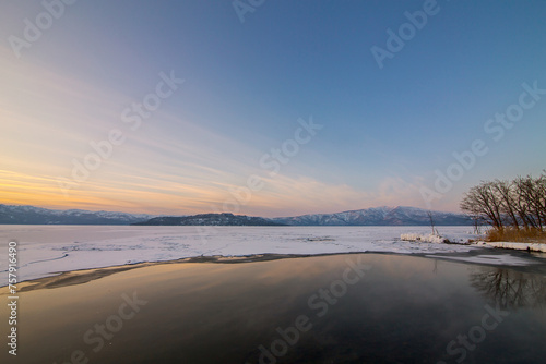 美しい夕暮れの空と湖畔の森を水面に反射する凍った湖。北海道の屈斜路湖。