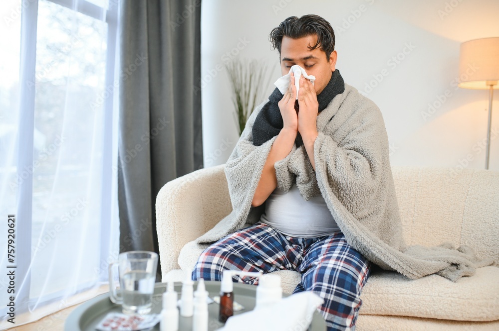 Sick man sitting at home on sofa in living room, sneezing has allergy and runny nose, Indian man with napkin has a cold Sick man sitting at home on sofa in living room, sneezing has allergy and runny nose, Indian man with napkin has a cold