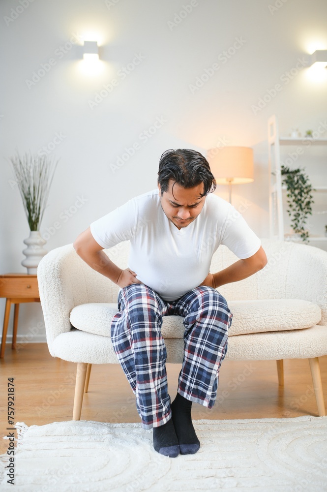Unhappy indian or arabian man, sits on comfortable sofa in cozy living room, holds his hands on his stomach, grimaces from pain in his stomach, suffers from poisoning, spasm, stomach problems.