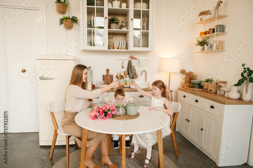 Happy mother with her daughter and son drinking tea or cocoa in the kitchen and having fun. Family communication of mom and children. Children congratulated their mother with flowers on Mother's Day.