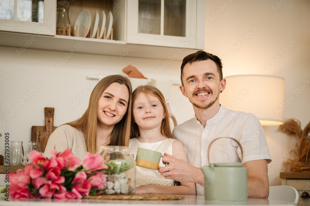 Family communication and spending time together. Mom and Dad hug a child. Mommy and Daddy embrace children. Mother, father, and daughter drinking tea or cocoa sitting in the kitchen, and having fun. Family communication and spending time together. Mom and Dad hug a child. Mommy and Daddy embrace children. Mother, father, and daughter drinking tea or cocoa sitting in the kitchen, and having fun.