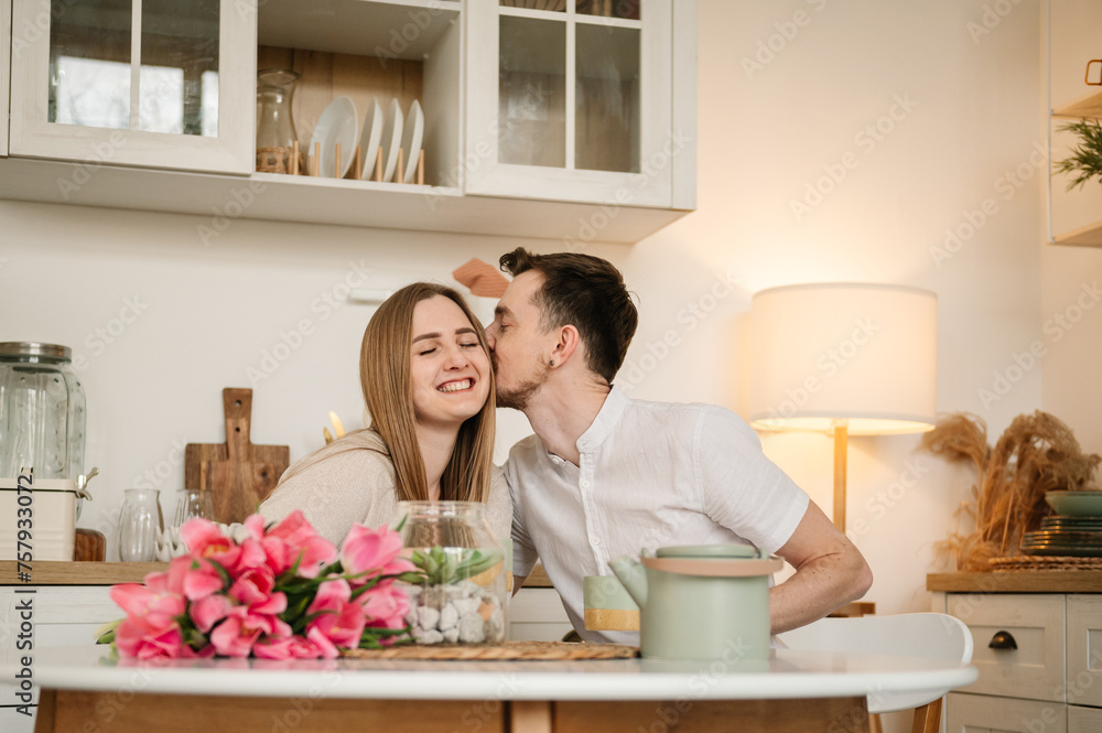 Romantic male kisses female, spending time together. Loving young couple having conversation and drink tea in morning. Couple drinking coffee in the kitchen. Man congratulate woman on women's day.