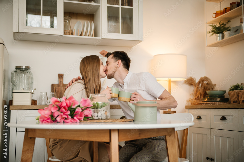 Romantic female and male kissing, spending time together at home. Loving young man and woman having conversation and drink tea in morning. Couple drinking coffee, holding cups in hands in the kitchen.