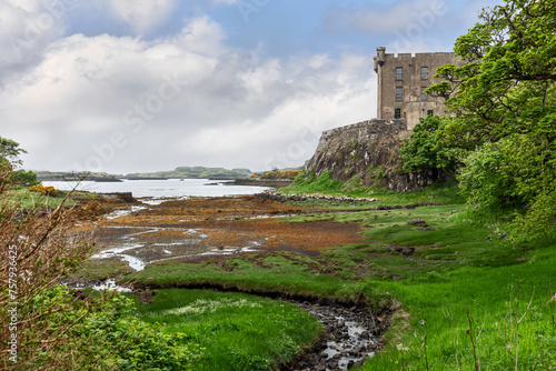 The historic Dunvegan Castle on the Isle of Skye, framed by vibrant green flora and a pebbled shoreline, under a dynamic sky, evoking Scotland's rich heritage photo