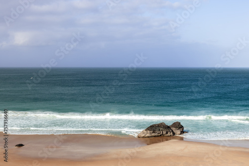 A serene view of Durness Beach, showcasing the tranquil blue waters of the Atlantic meeting the golden sands. A solitary rock formation stands near the shoreline, under a vast, partly cloudy sky photo