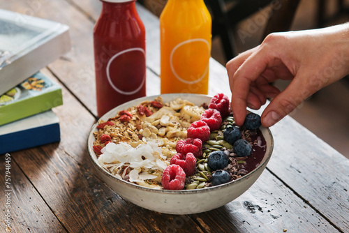 Female hands pick up a blueberry  from a Smoothie açai bowl with banana, strawberry, coconut flakes, chia, nuts, beautifully decorated. Served with bottle of juice on a wooden table.  photo
